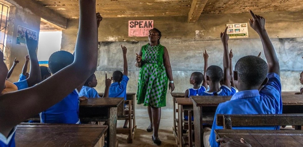 A teacher stands at the front of a classroom in Africa. Pupils sit in rows of wooden desks with their hands in the air.