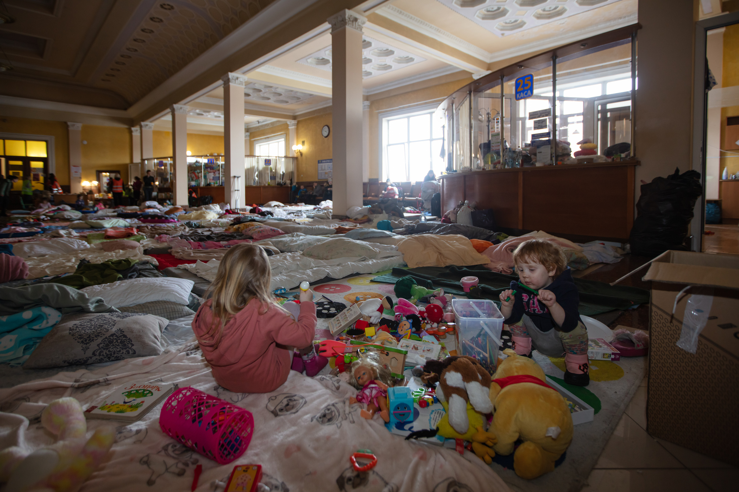 ukrainian children sit and play with toys on the floor of a railway station with sleeping bags across the floor