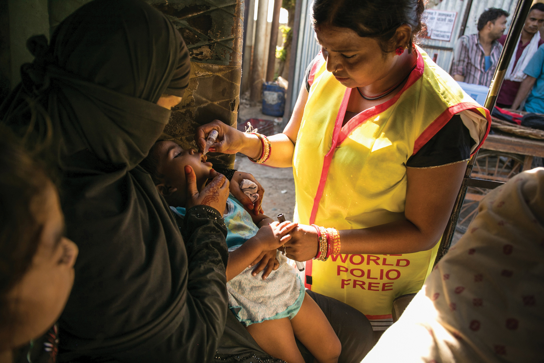 a person in a yellow End Polio Now vest administers the drops of polio vaccine into the mouth of a young child. India.