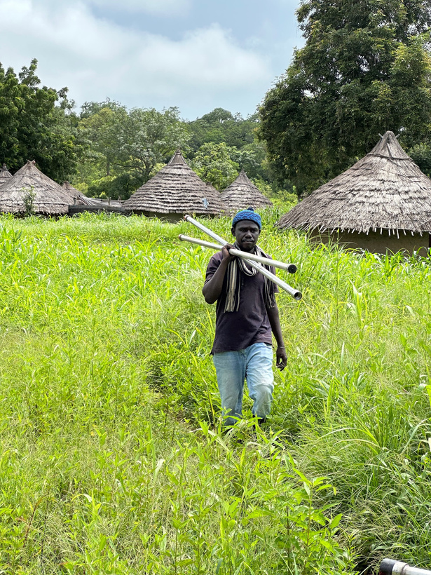 A person walking through a grassy field with thatched huts in the background.