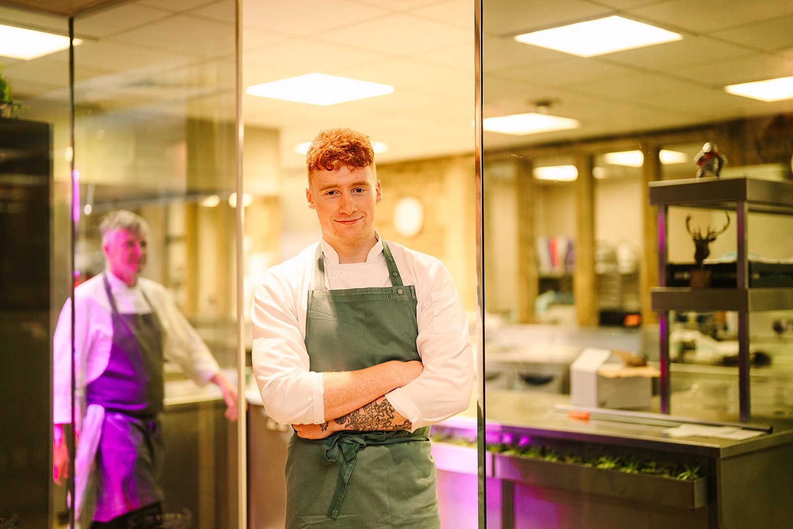 Tom Hamblet in an apron is standing in front of a glass door and smiling at the camera