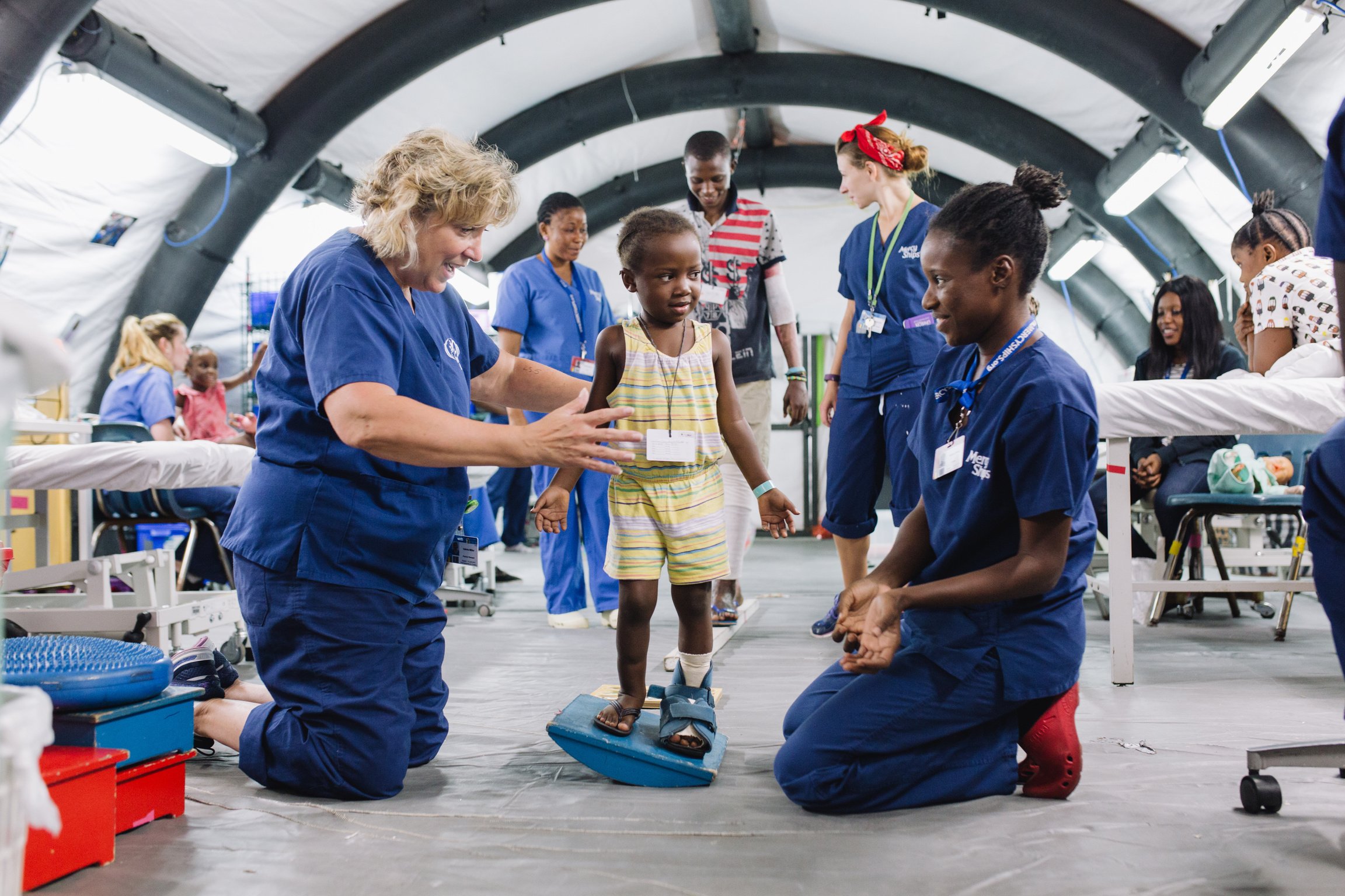 Two nurses help a young girl with her balance as she stands with a medical boot on her foot.