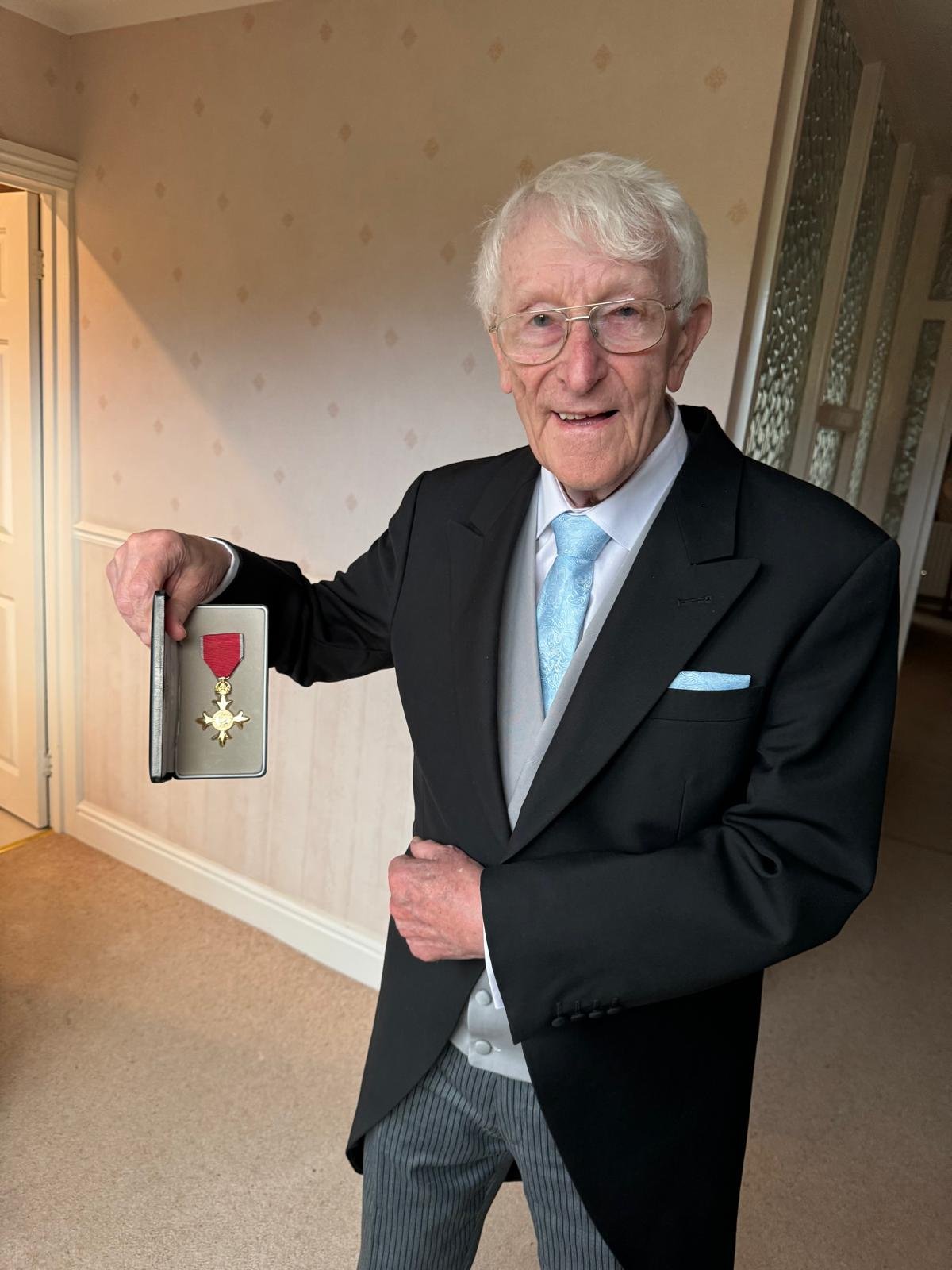 Rotarian Paul Denton stands in a suit holding his OBE medal in a presentation box