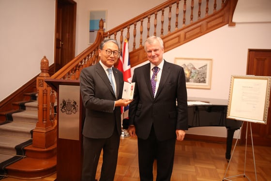 Two individuals in suits and ties standing next to each other, one holds a small award with a British flag in the background.