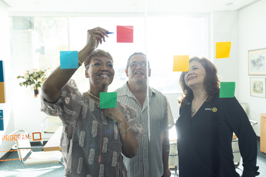 A group of three Rotary members plan a project by arranging sticky notes on a glass wall.