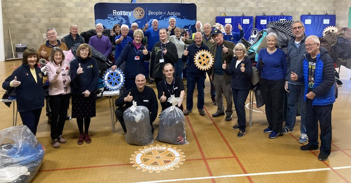 A group of Rotary volunteers in a gym posing for a picture with bags of donated clothing and cut-outs of the Rotary logo