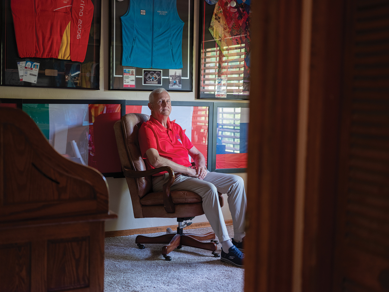 an olympic volunteer sits in his office with framed flags and volunteer uniforms