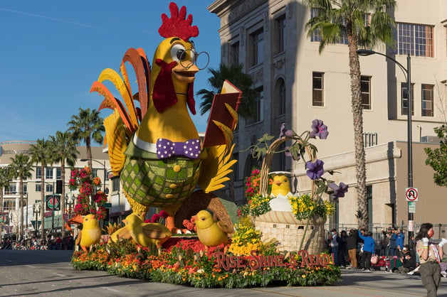 A large float with a chicken wearing glasses on it in the middle of a street at the Rose Parade.