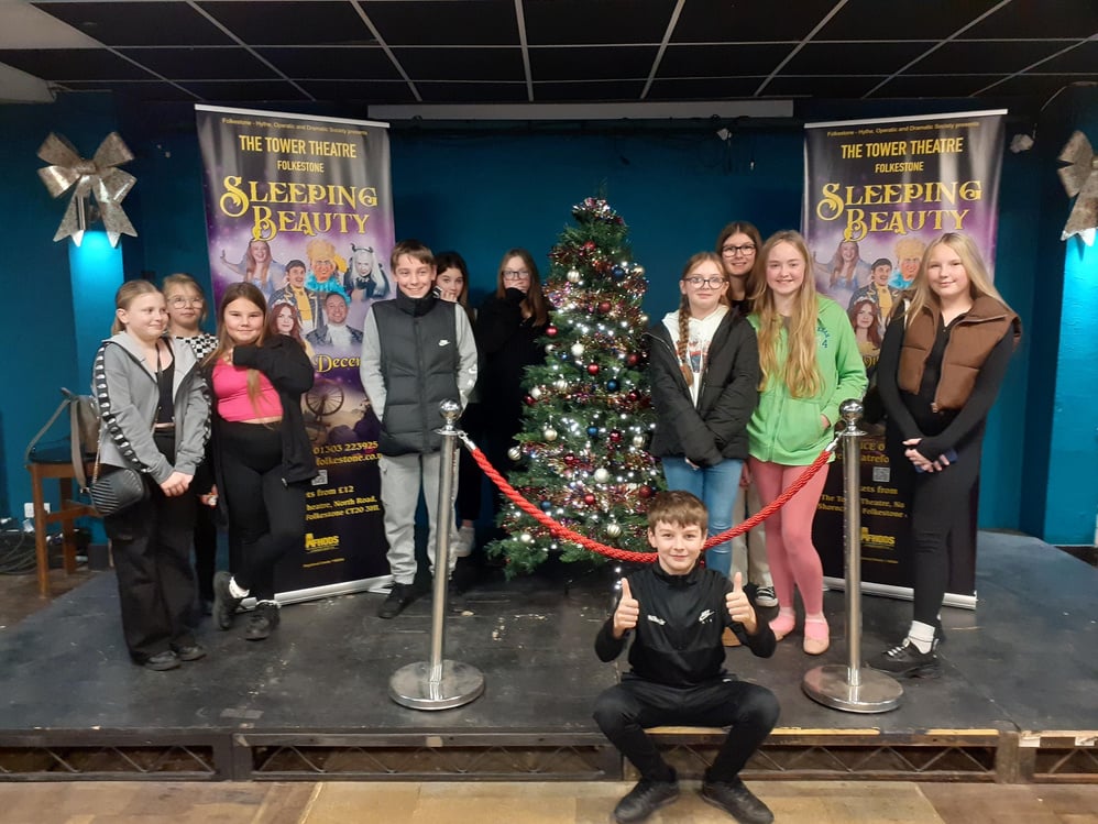 A group of young people posing in front of a Christmas tree and advertisements for a production of 'Sleeping Beauty'.