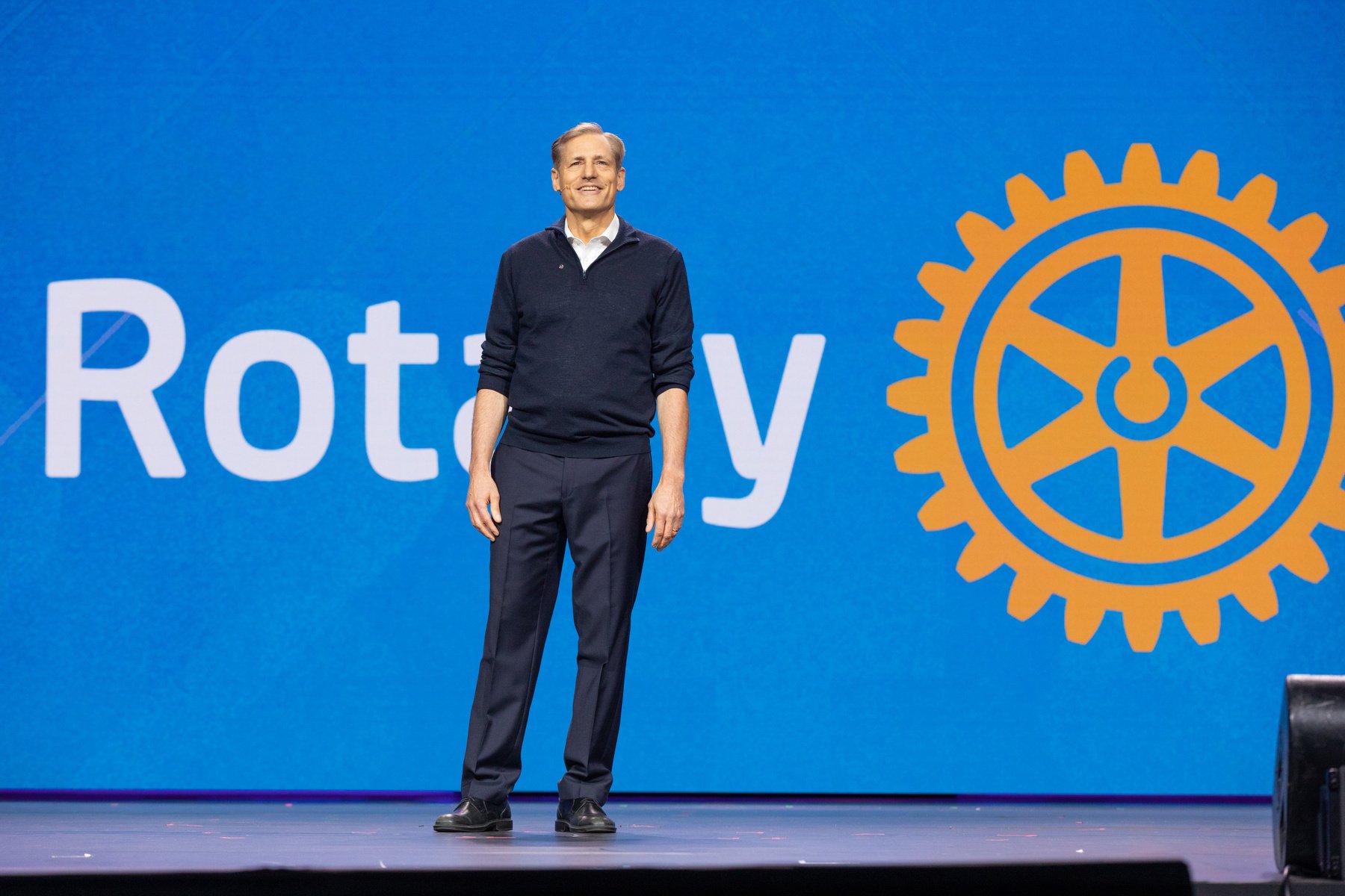 John Hewko stood on stage with a large Rotary logo on a big screen
