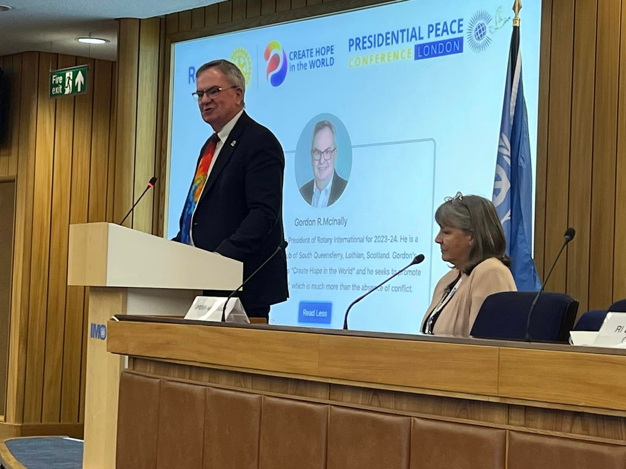 RI President Gordon McInally standing at a podium in front of a large screen at the Presidential Peace Conference.