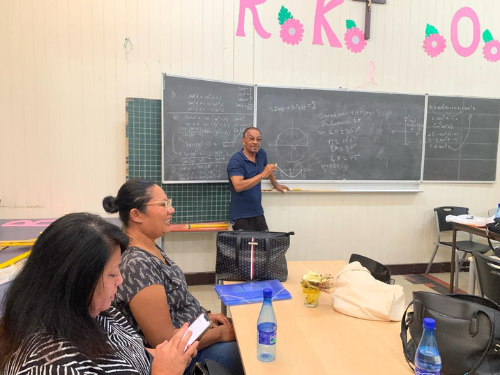 a person holding chalk in front of a blackboard whilst talking to a classroom full of people