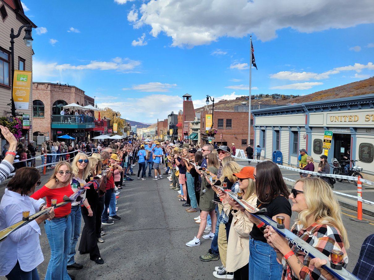 A long line of people standing in front of a building whilst holding skis balancing shot glasses.