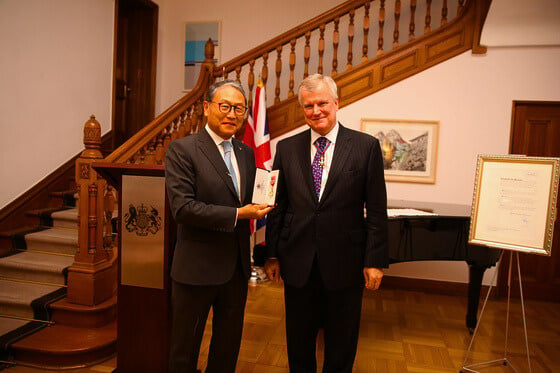 two people in suits and ties standing next to each other. One holds an award with the British flag in the background.