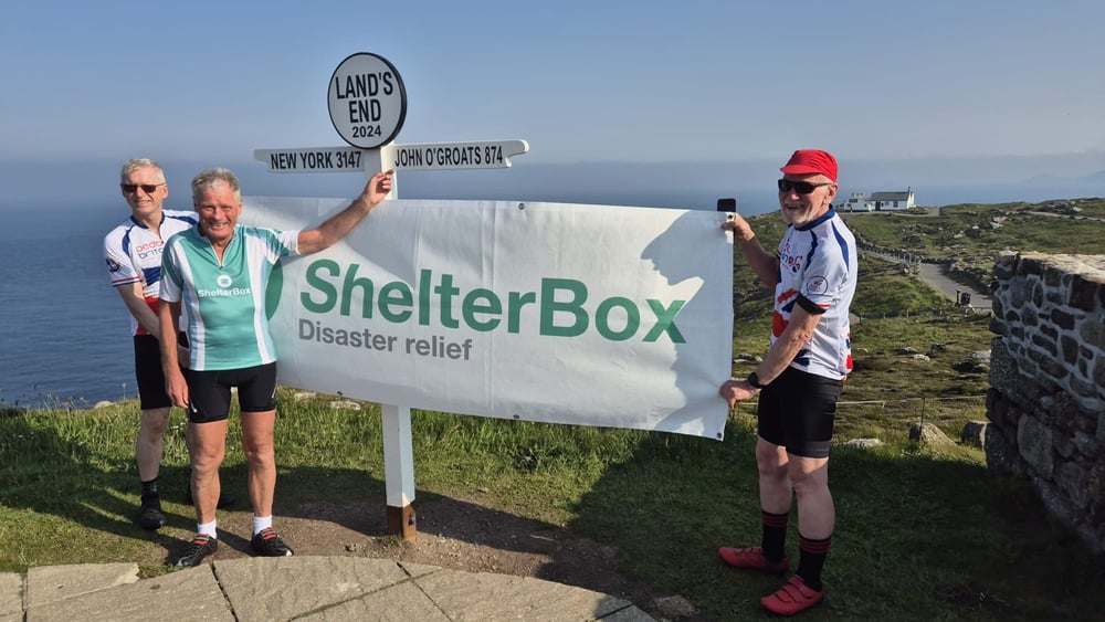 three people pose for a photo in front of a sign that says shelterbox