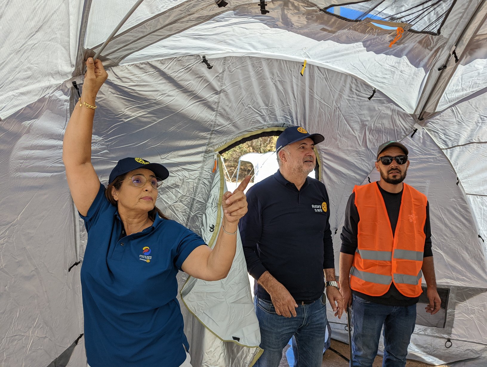 Two people in Rotary caps and one in sunglasses and high-vis standing inside a ShelterBox tent.