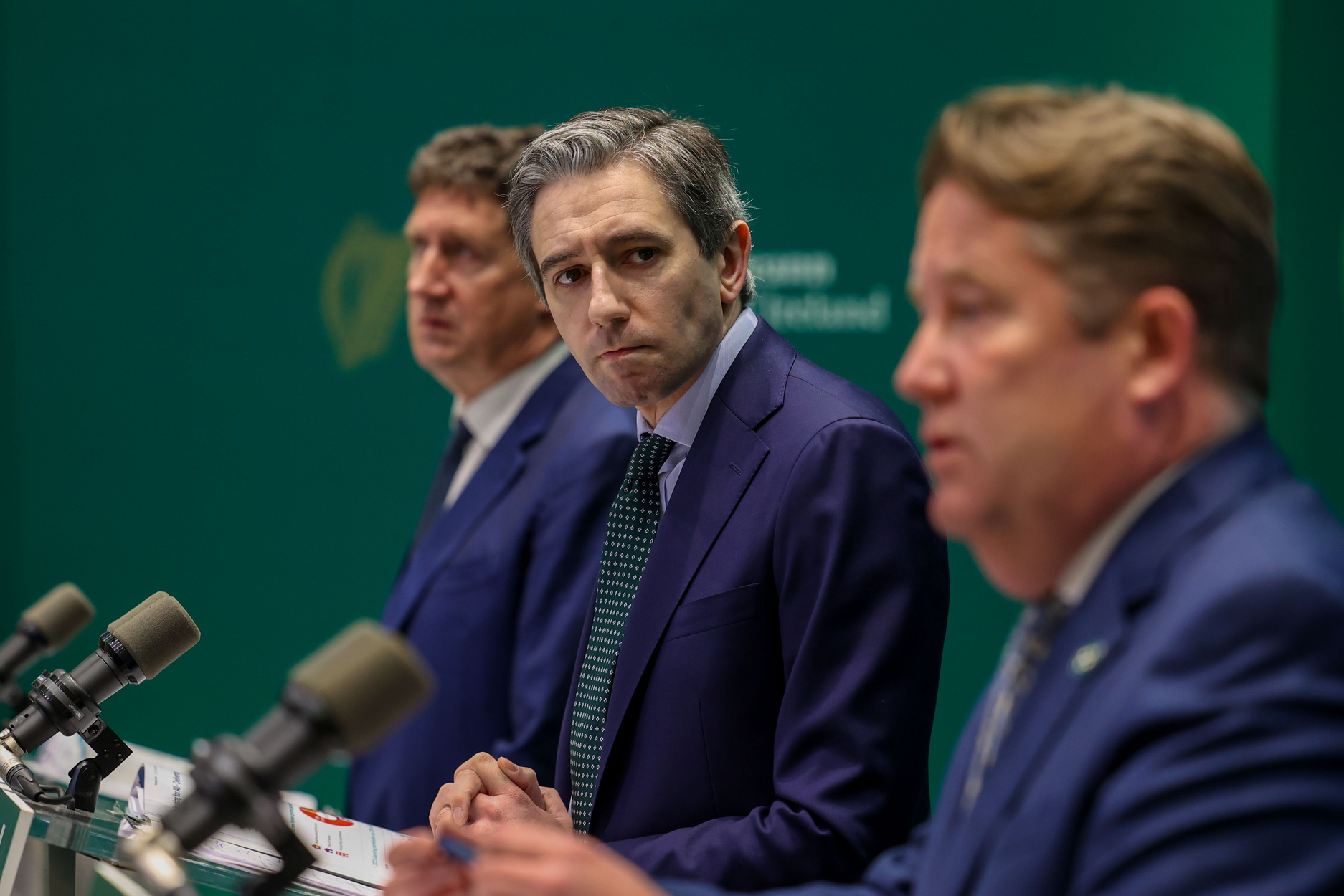 Simon Harris standing at a press conference podium with two other blurred men