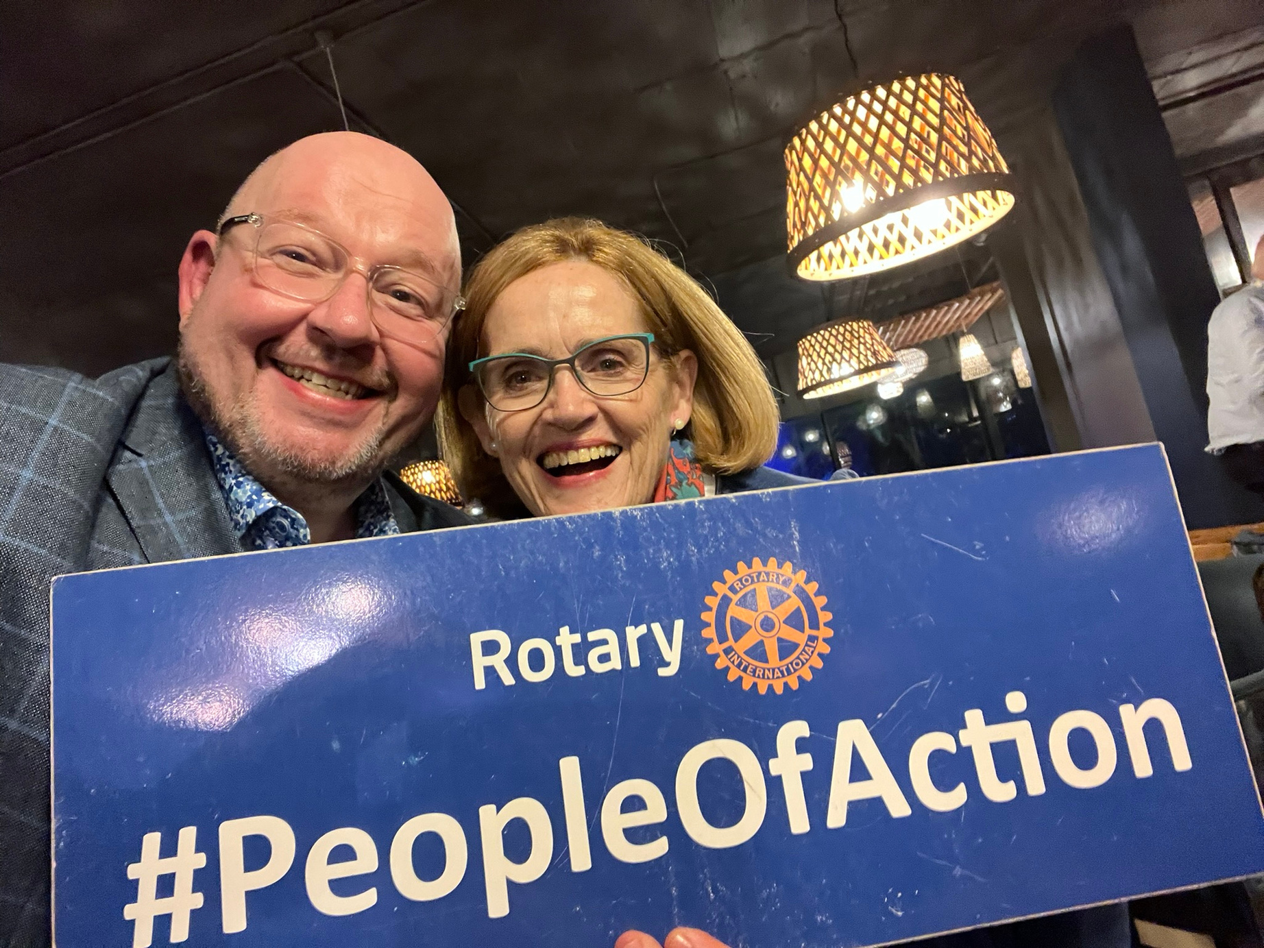 Two Rotarians pose for a selfie with a Rotary sign reading #PeopleOfAction