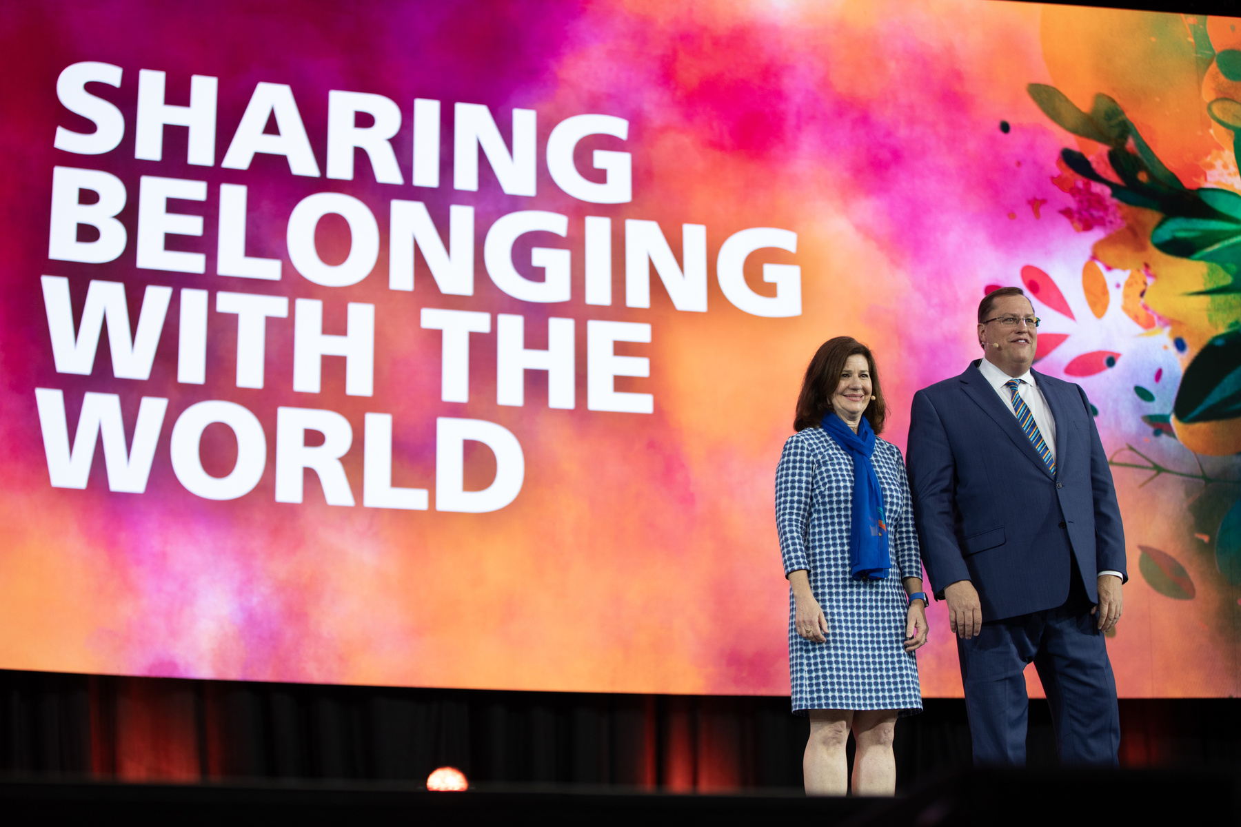 Tom Gump and his wife stood on stage at RI Convention with the words 'Sharing Belonging with the World' on the big screen.