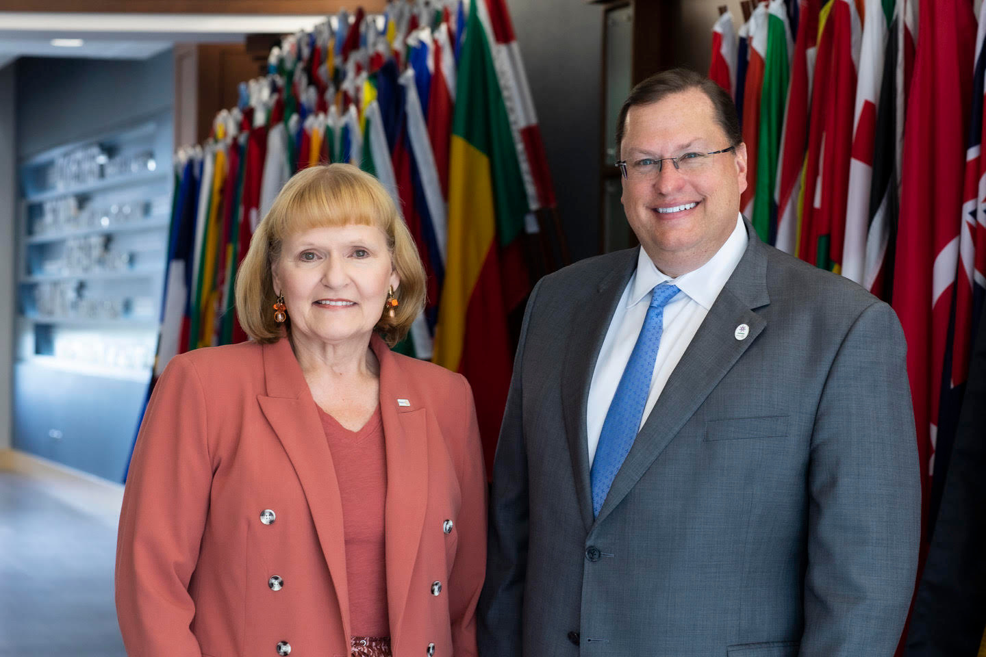 RI President Stephanie Urchick and her aide Tom Gump stand smiling in front of flags of the world.