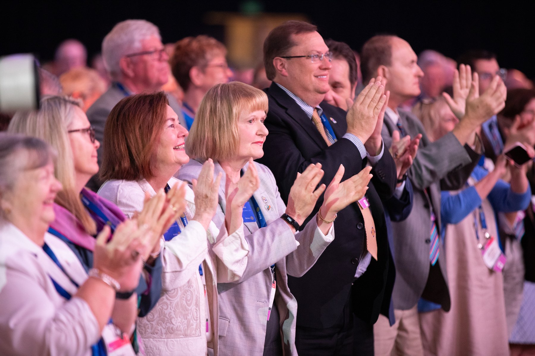 Tom Gump and Stephanie Urchick stand in a crowd applauding in a Convention auditorium. 