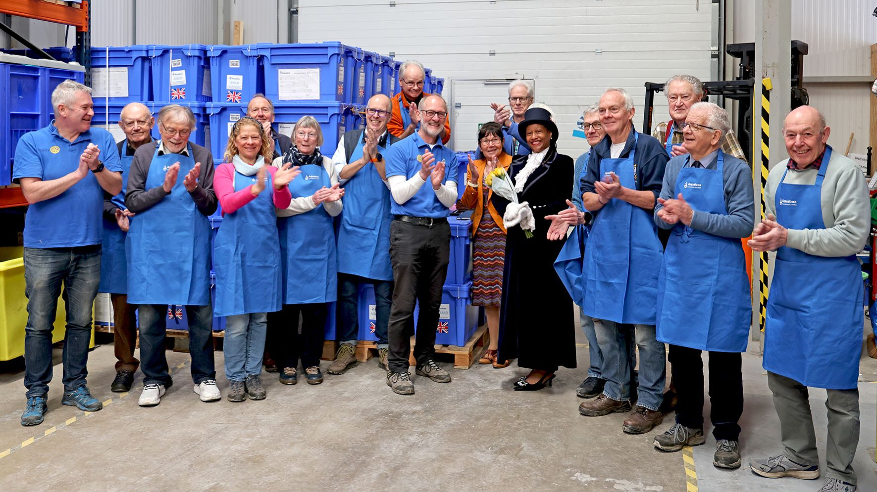 Aquabox volunteers stand smiling and applauding in front of stacks of blue Aquabox crates