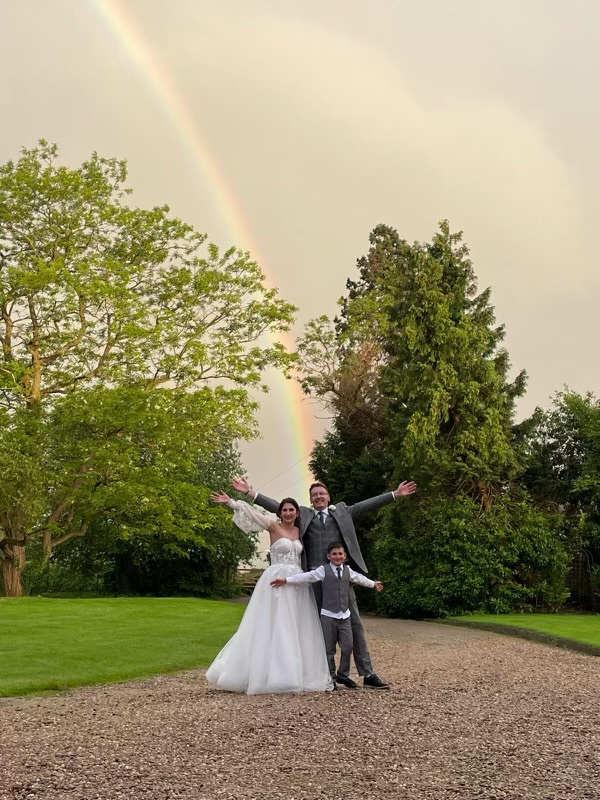 a bride and groom pose with their arms outstretched in front of a rainbow