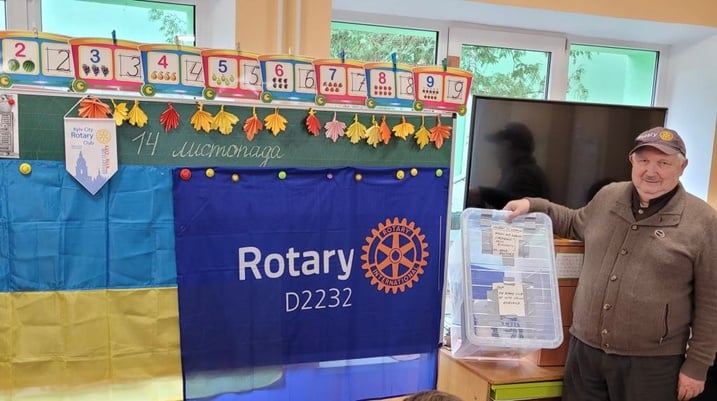 Rotarian Sergii Zavadskyi in a classroom in front of a Rotary banner holding a friendship box sent by Rotary Great Britain & Ireland.