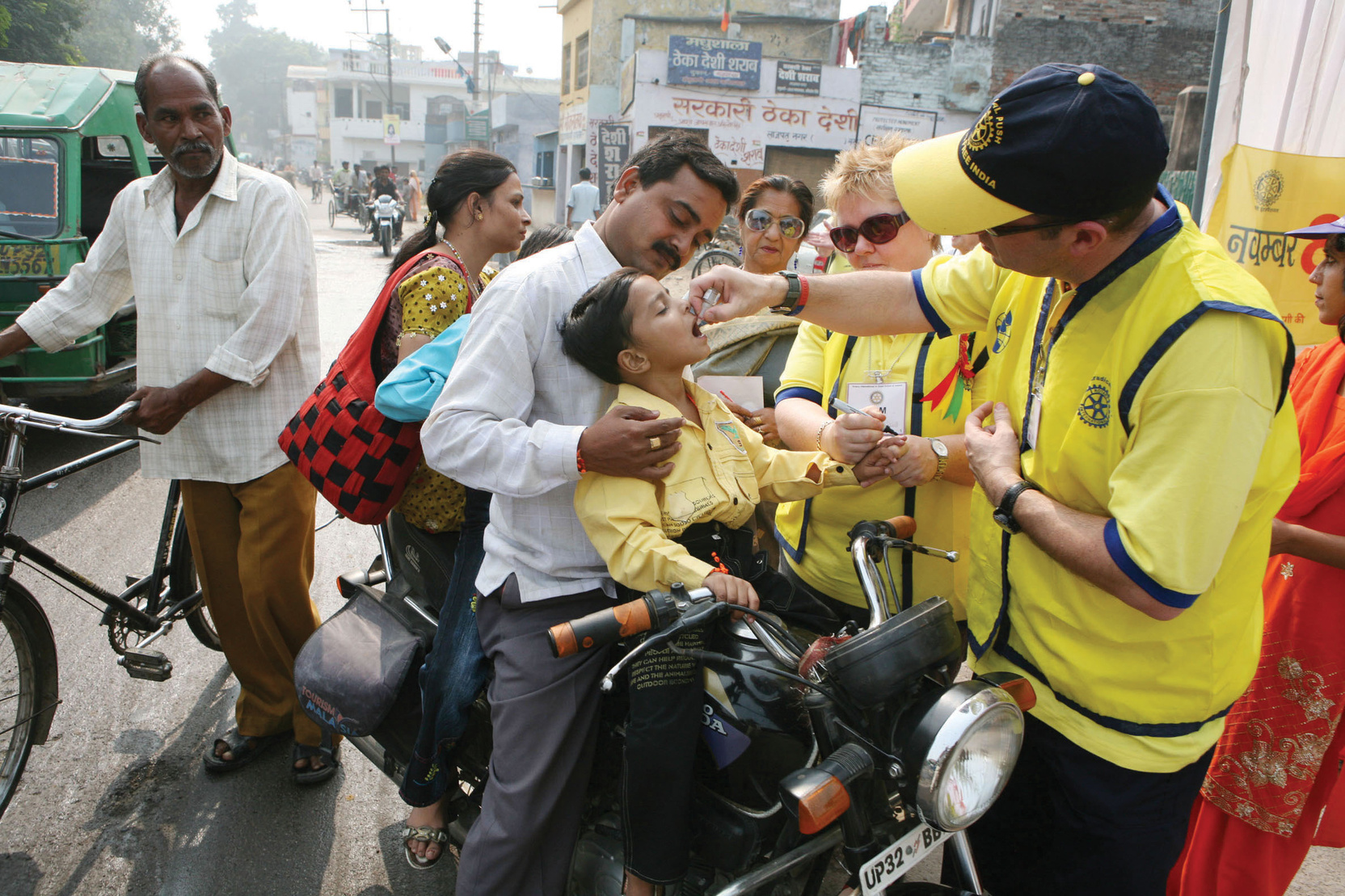 A polio worker in a yellow giving a child on a motorcycle a polio vaccination